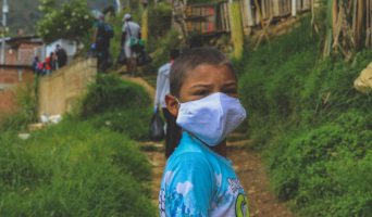 a young child walking on a path outside, turned around to view the camera wearing a mask