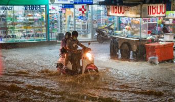 heaving raining causing flooding on a street in Thailand, with two people trying to drive through on their scooter