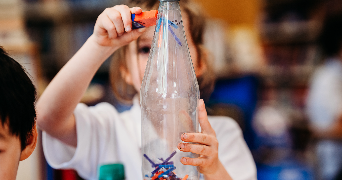 an image of a child in the classroom displaying how magnets work by using magnetic objects in a plastic bottle and a magnet on the outside of the bottle