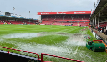 Flooded football pitch with standing water near the sidelines and empty stadium seating in the background