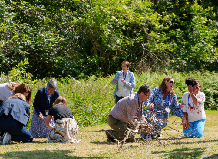 Groups of adults kneel and crouch on grass in an outdoor workshop setting, exploring natural materials like sticks and twigs. They appear engaged and collaborative, surrounded by greenery and trees on a sunny day