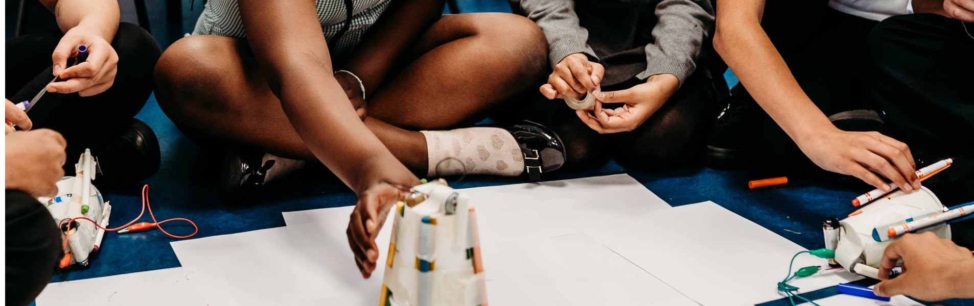 Children sitting on the floor working together on a hands-on science activity with paper, pens and small hand-made robotic creations
