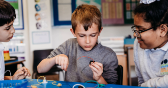 Three primary school children work together on a hands-on science activity, exploring electrical circuits with wires and a battery