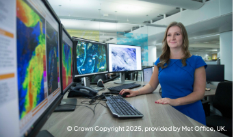 a scientist working at a desk with multiple computer monitors displaying weather maps and satellite imagery in a modern office/lab setting