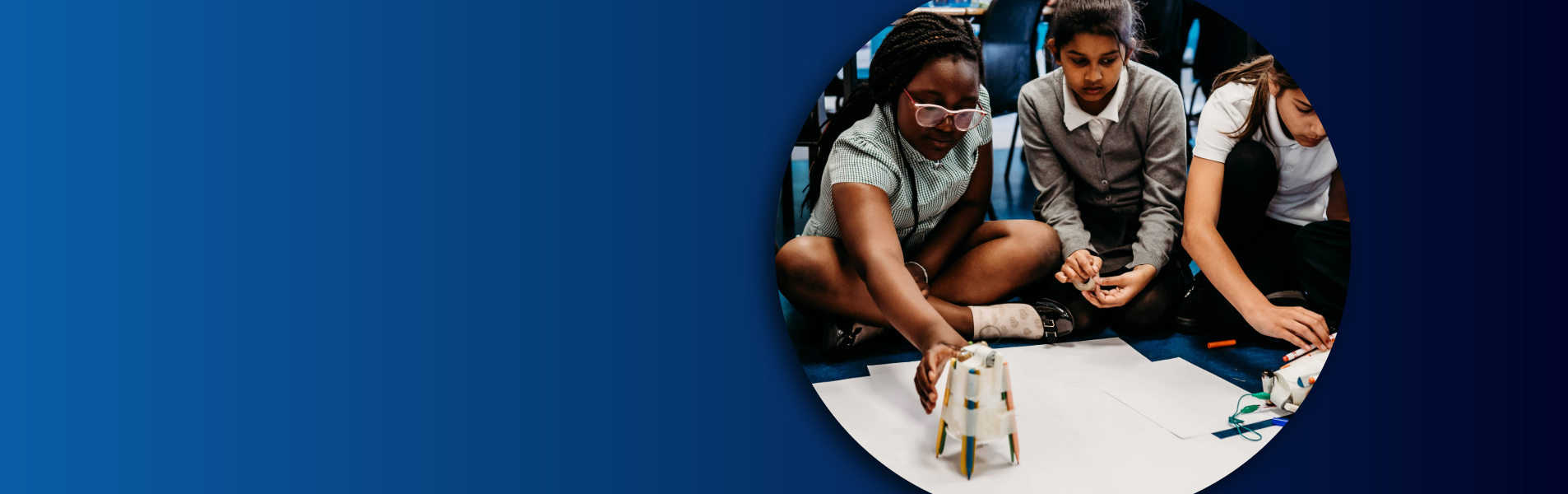 Three primary-aged pupils sitting on the floor, working together to build a small structure using recycled materials during a science activity