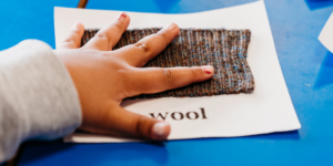 a child's hand touching a textured wool fabric sample during a practical science activity exploring materials