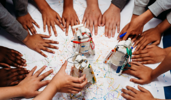 children’s hands surround a homemade robotic device using marker pens to create drawings on paper